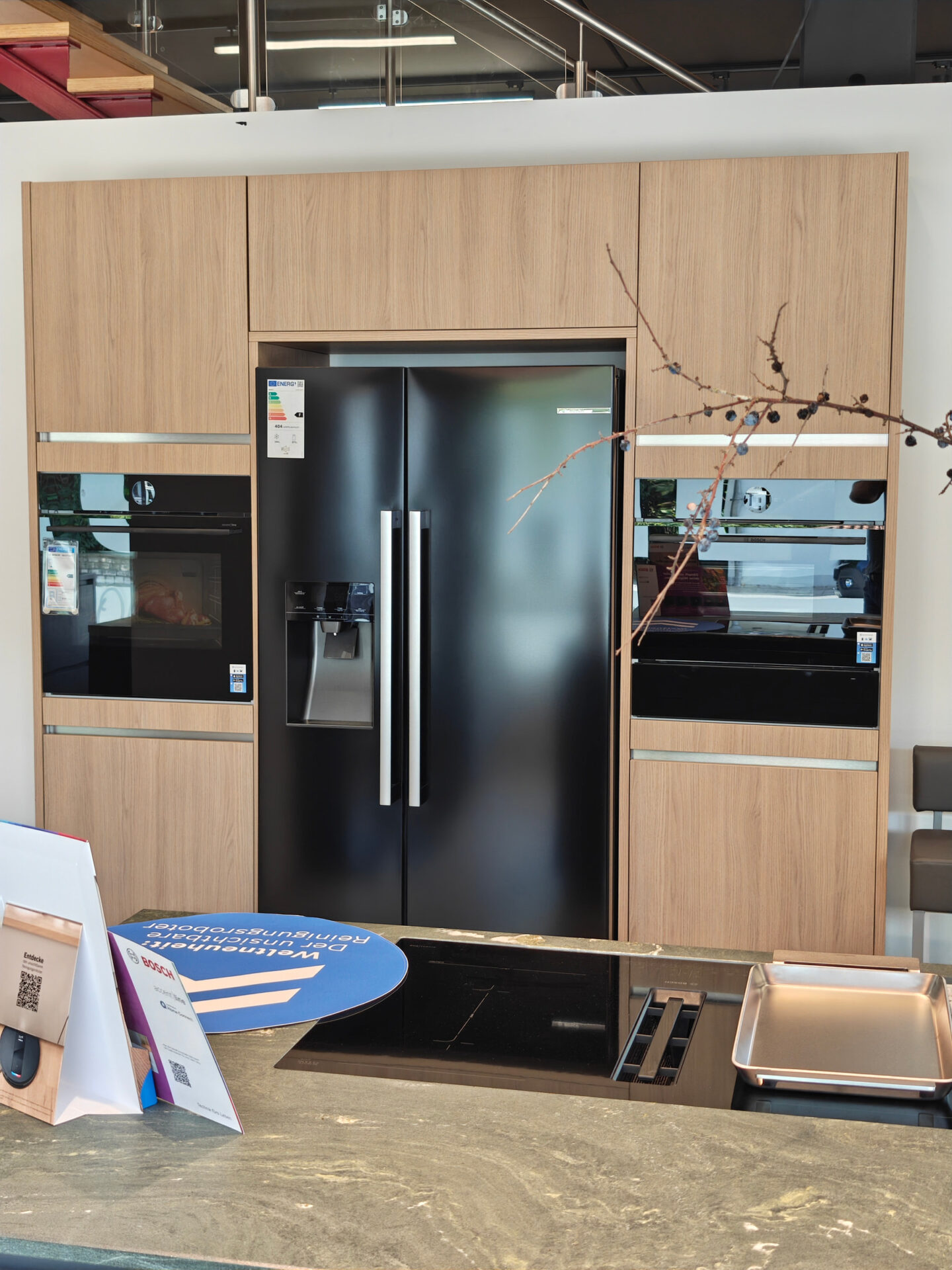 Hochschrankwand in Feineiche natur mit zwei Einbaugeräten und schwarzem Side-by-Side-Kühlschrank mit Wasserspender.